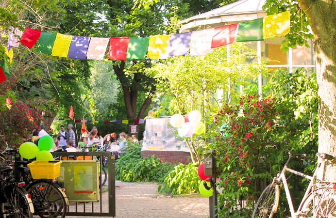 Foto. Der Eingang zum Stadtteilzentrum. Über dem Gartentor wehen bunte Wimpel, im Garten stehen und sitzen feiernde Nachbar*innen.